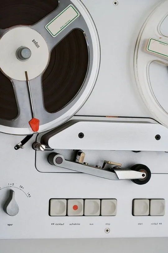 Close-crop of a white reel-to-reel deck, soft grey keycaps and a single red accent against brushed aluminium, a study in restrained control-surface design.